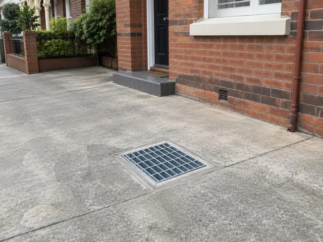 Dry outdoor concrete driveway with a square metal drain grate beside a brick house in Annandale