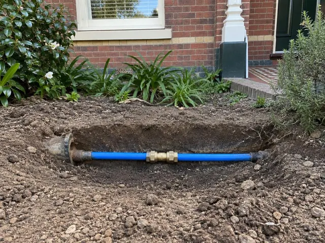 Exposed blue water pipe with brass coupling in a front yard trench beside a brick house in Camperdown