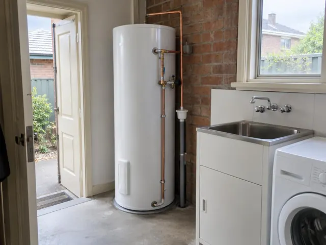 Hot water cylinder beside a sink and laundry area with brick wall and open door in Stanmore